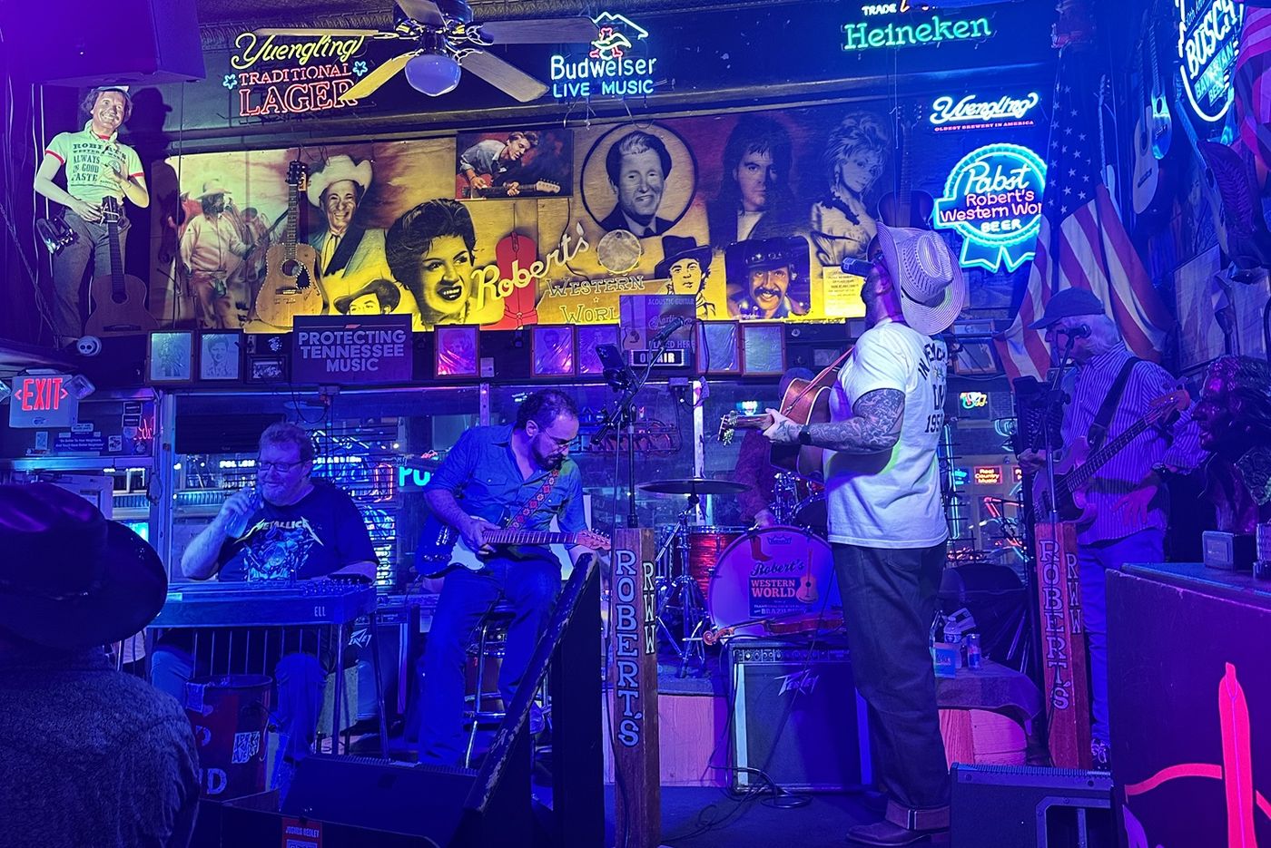 A band playing on the stage at Robert's Western World, with a steel guitar on the left, under a wall of neon signs and a mural of country legends